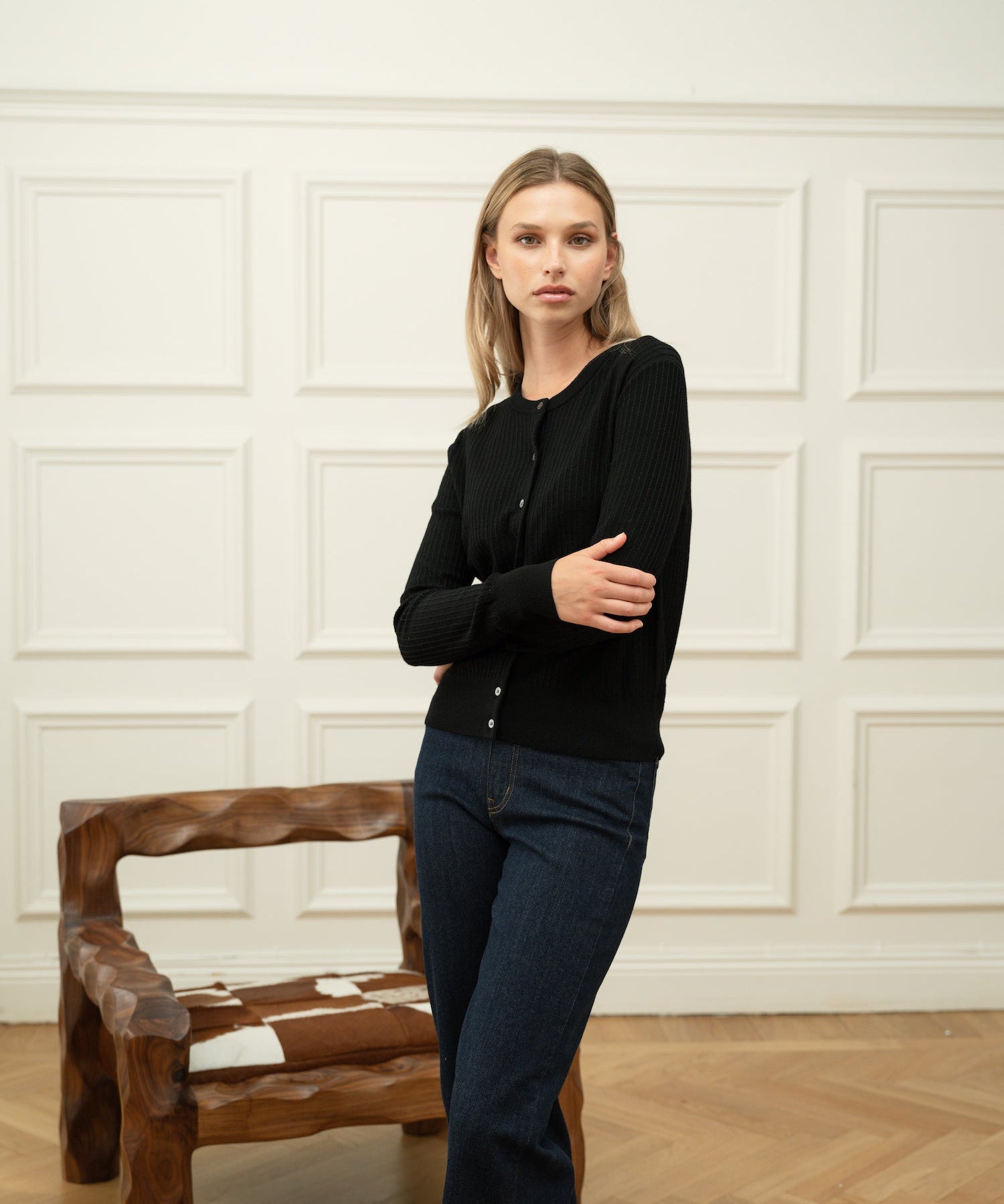 Woman wearing a Ninepine black merino wool cardigan with dark blue denim jeans against an off-white wainscoted wall